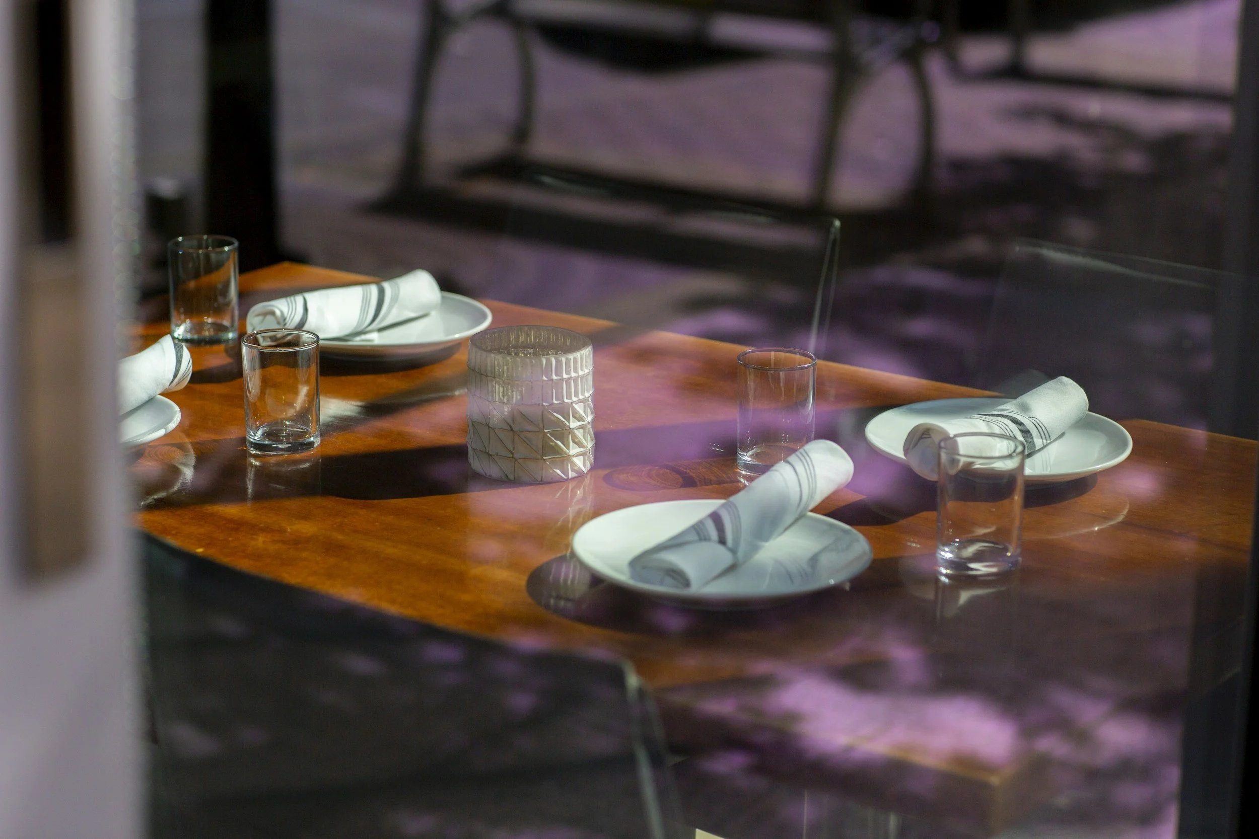 Table setting with four plates, four rolled napkins, four glasses, and a decorative jar, seen through a glass window.