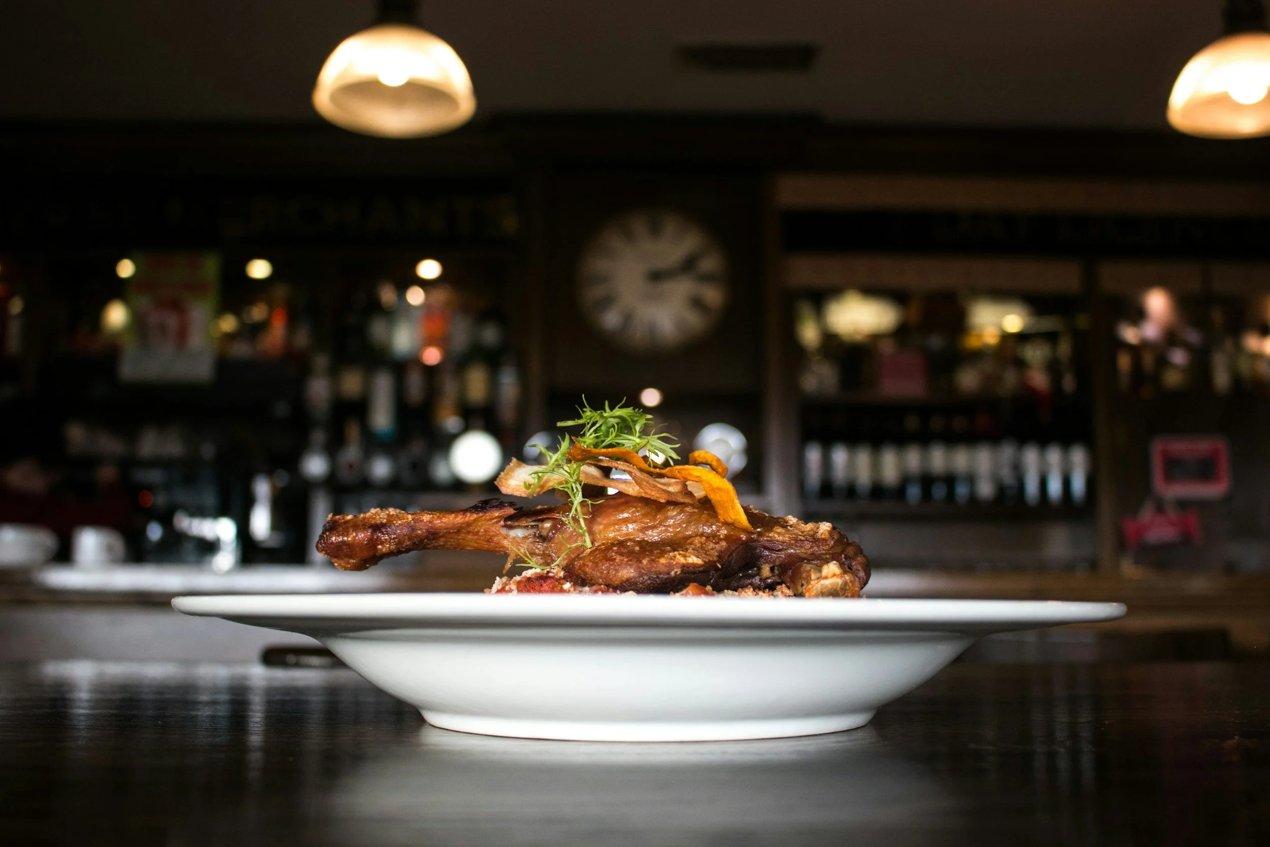 Close-up of a plated cooked meat dish garnished with herbs and vegetables, in a dimly lit restaurant or bar setting.