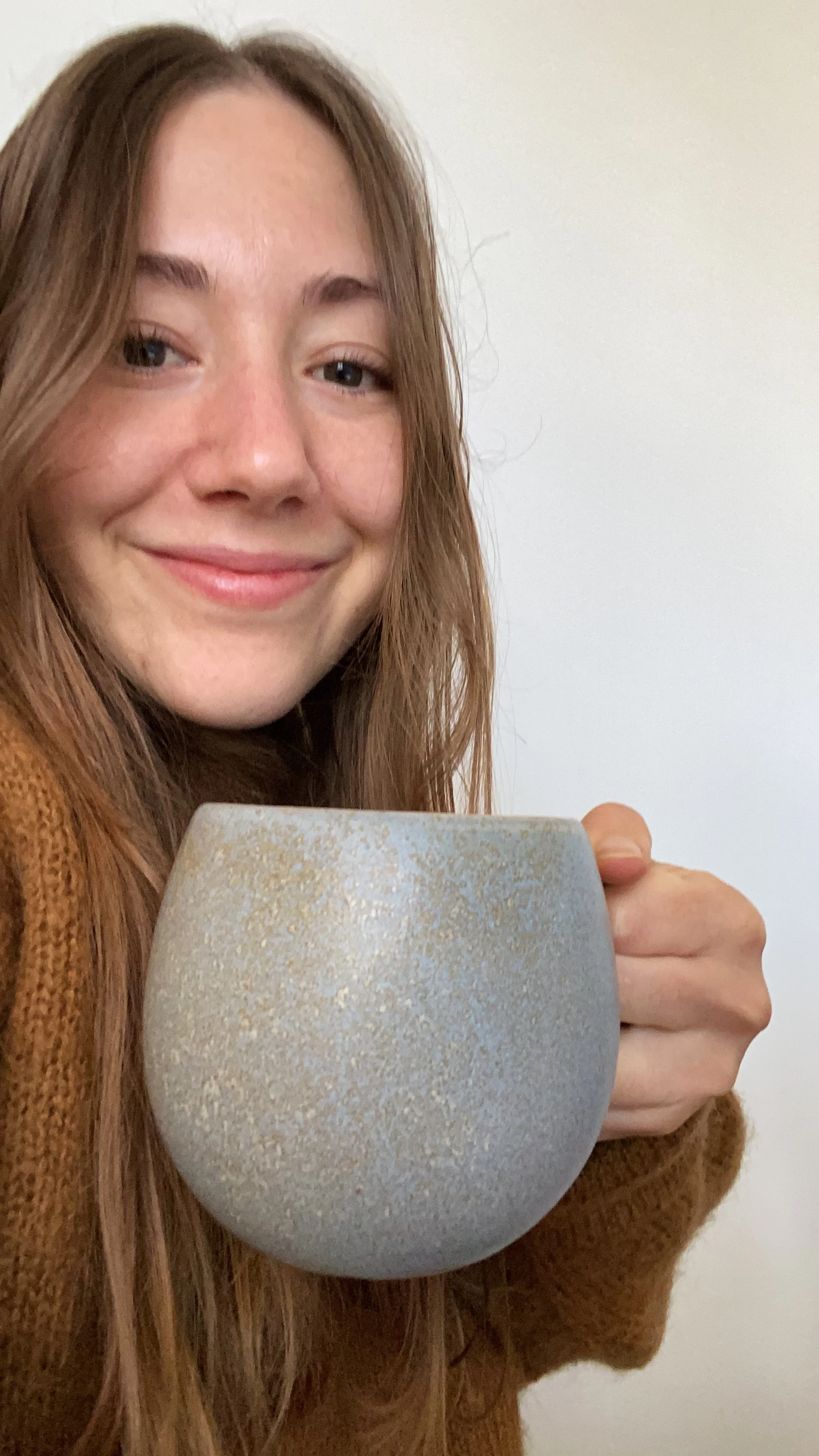 A young woman with long, light brown hair smiling while holding a large ceramic mug.