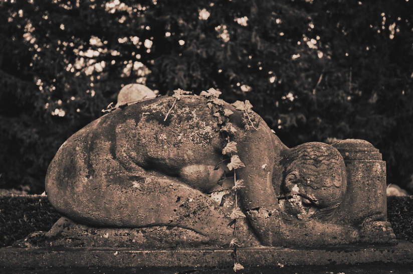 Weathered stone sculpture of a sleeping child, adorned with vines, set against trees in the background.
