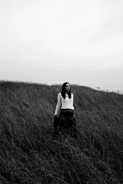 A woman standing in tall grass on a hillside, looking into the distance, in black and white.