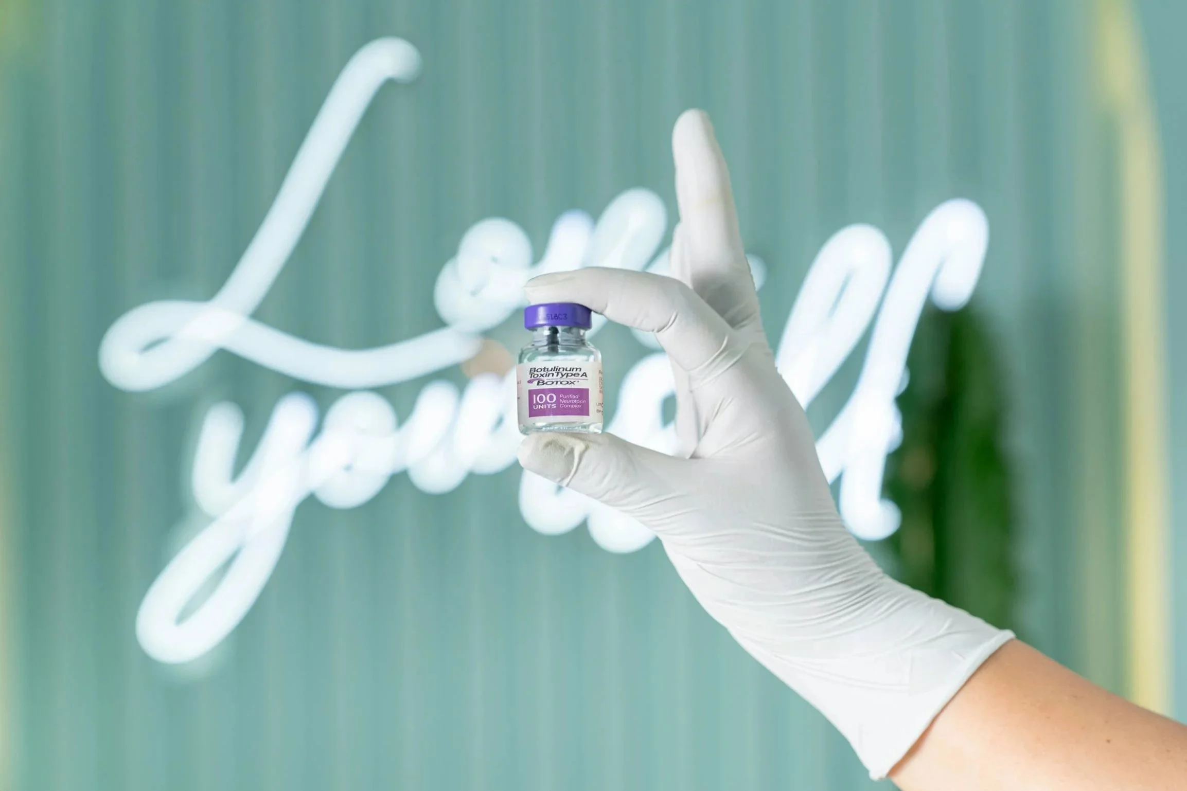 Hand in einer weißen Latexhandschuh hält eine kleine Vial mit einem Impfstoff, im Hintergrund leuchtet eine Neonbeschriftung 'You got this'.