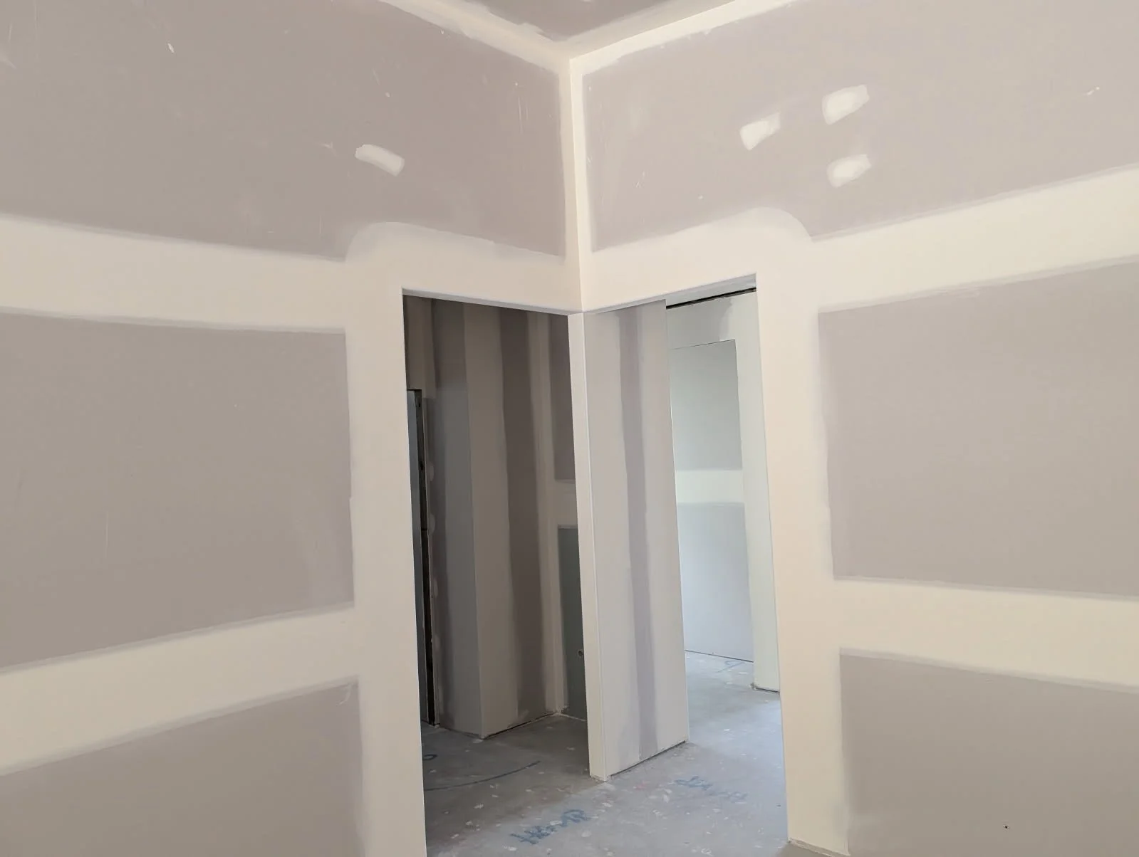 Interior view of a room under construction, showing drywall walls and ceiling, with visible joints and patches, and an open doorway leading to another unfinished room.