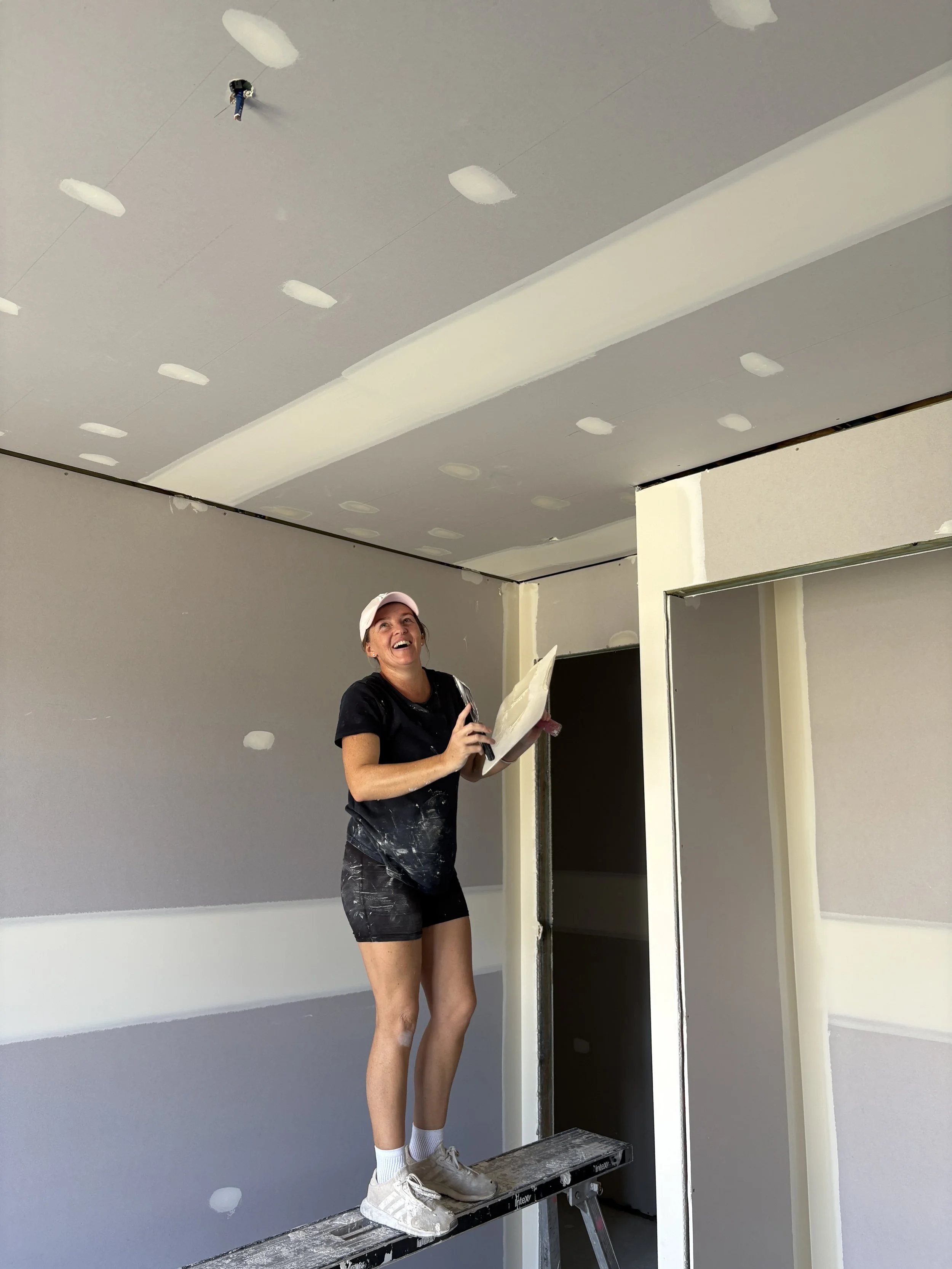 A woman standing on a ladder in a room under construction, holding a trowel and a plasterboard, smiling while working on drywall installation.