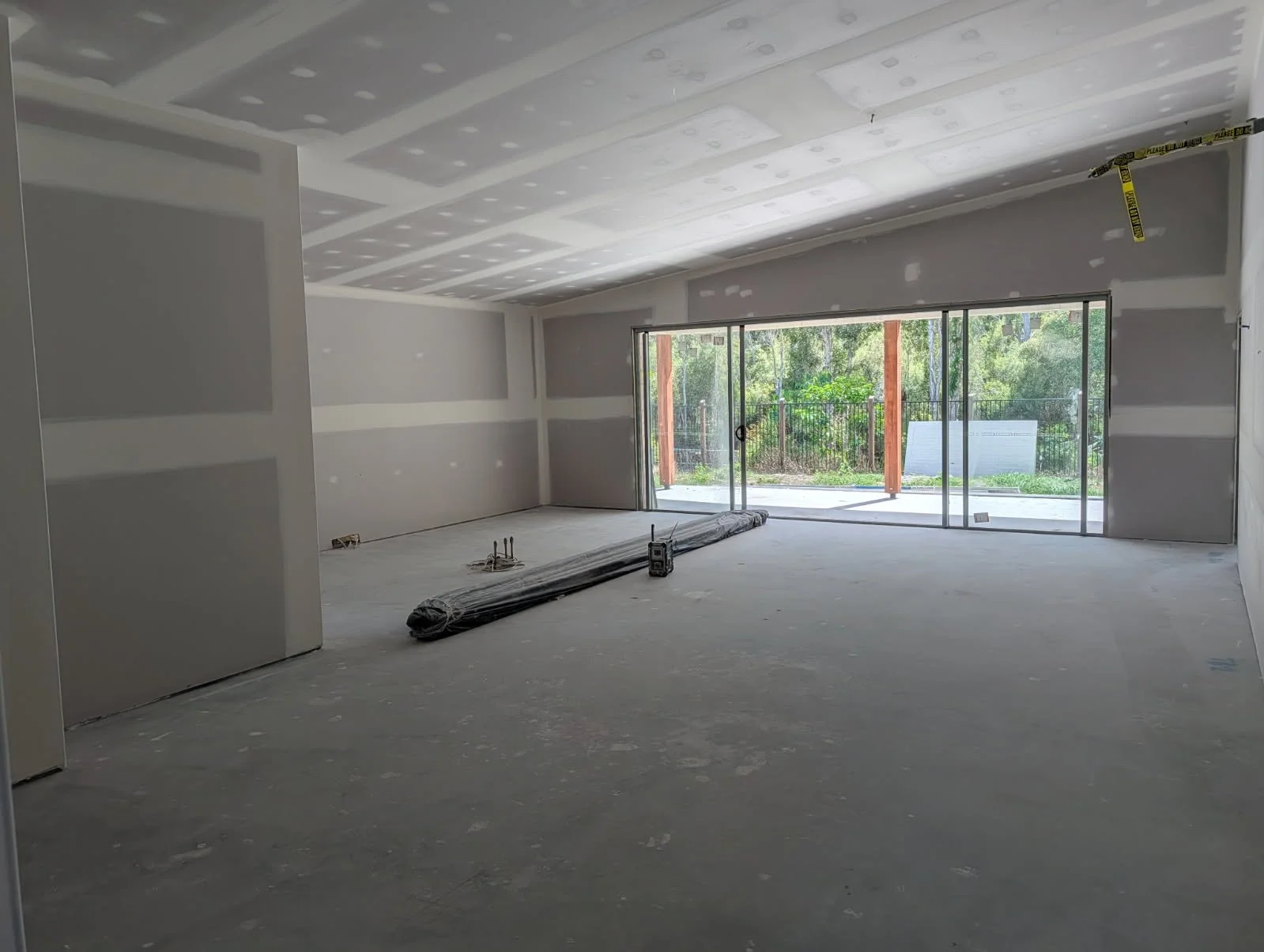 Interior of a house under construction with drywall walls and ceiling, large sliding glass doors leading to an outdoor patio, and construction materials on the unfinished floor.
