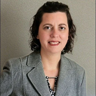 A woman with shoulder-length dark hair, wearing a gray blazer and a patterned blouse, smiling in front of a plain gray wall.