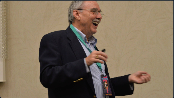 An older man with glasses, gray hair, and a beard, smiling and laughing, wearing a dark blazer, light blue shirt, and a lanyard with a badge, standing against a beige wall.