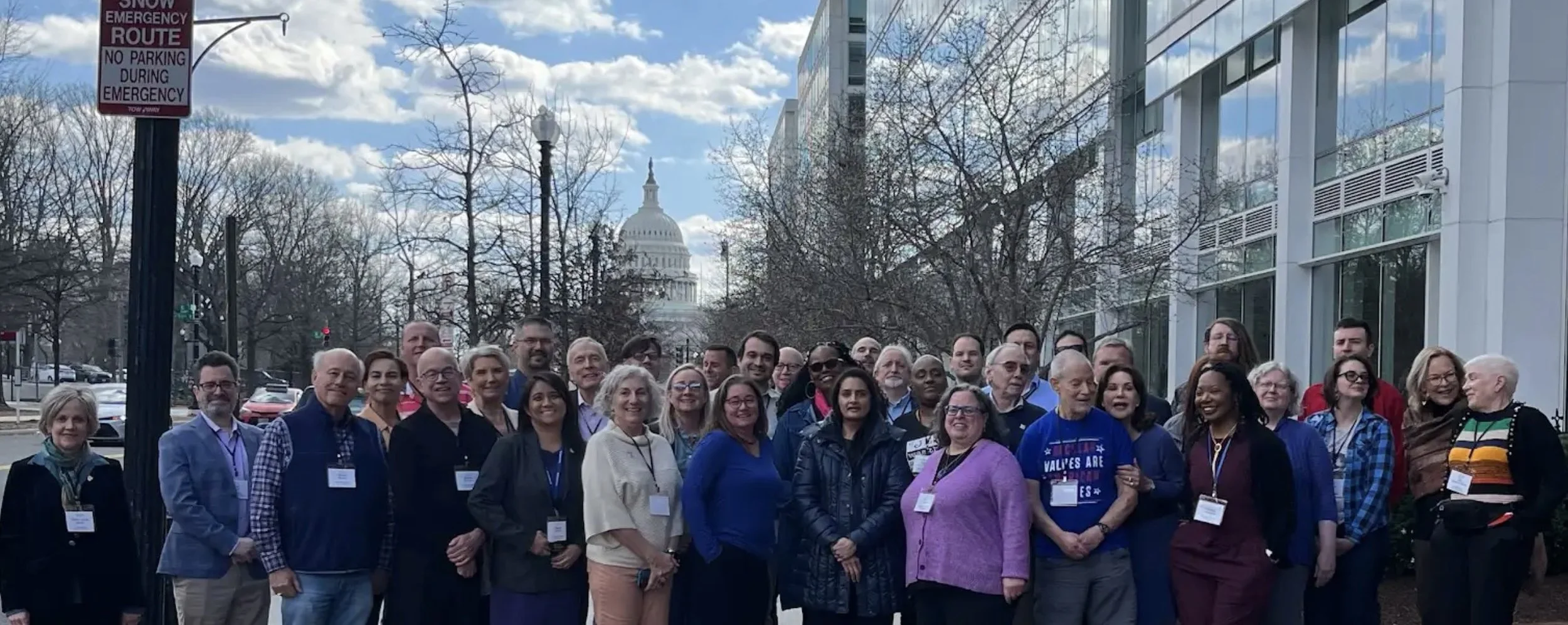 A group of diverse people poses for a photo outdoors in front of a modern glass building, with the United States Capitol building visible in the background. They are smiling and wearing name badges, suggesting a conference or gathering.