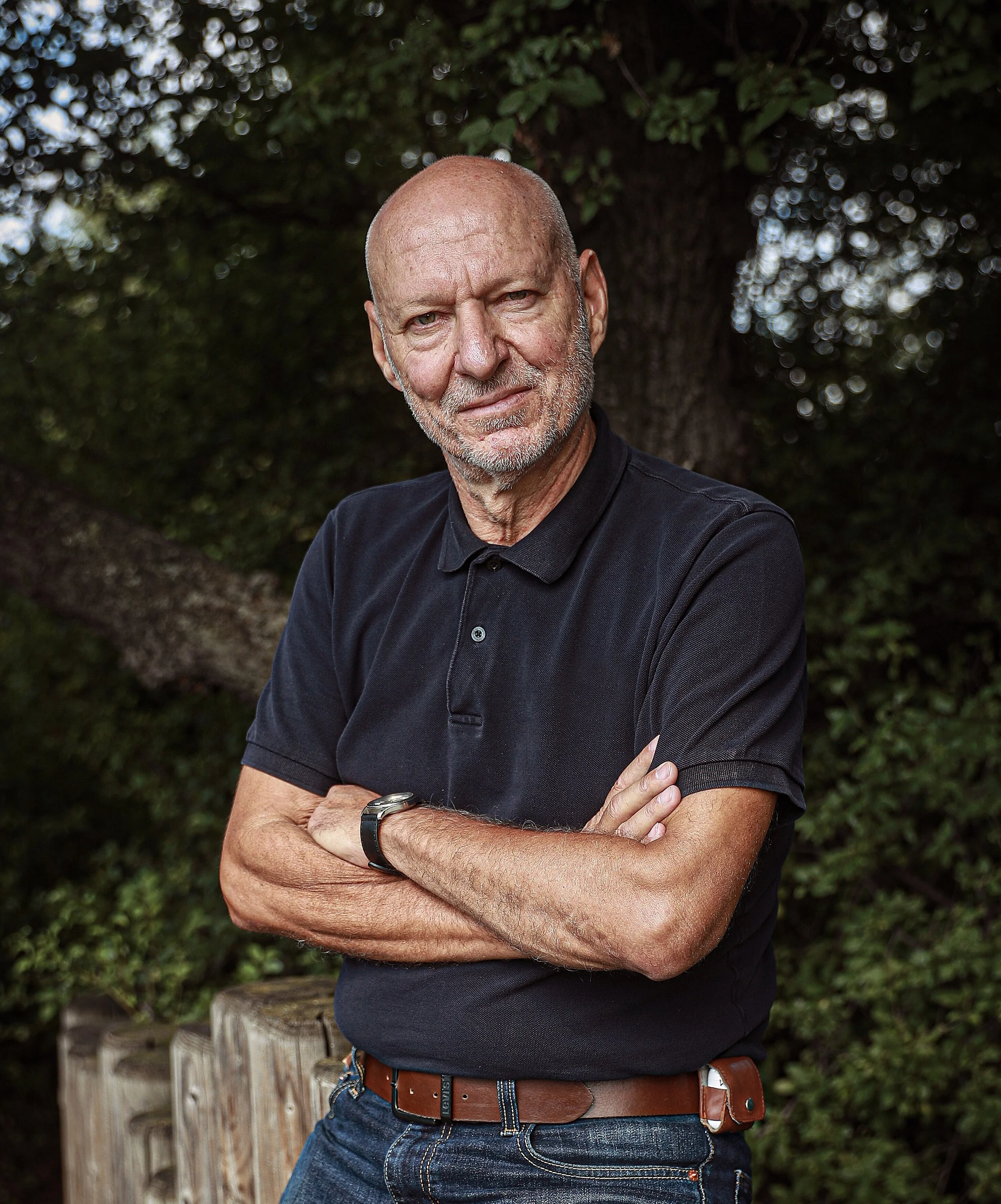 An older man with a bald head and gray beard stands outdoors with his arms crossed in front of a large tree, wearing a black polo shirt and blue jeans.