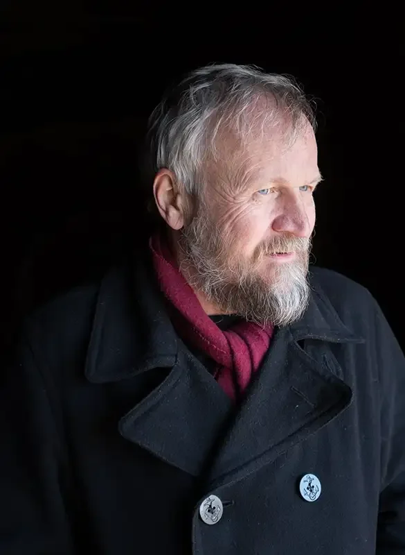 Profile of an older bearded man with gray hair, wearing a black coat and a red scarf, looking to the right against a black background.