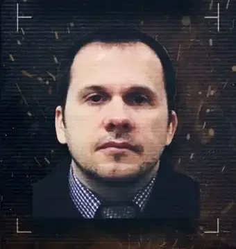 A portrait of a man with short dark hair, wearing a dark blazer and a checkered shirt, set against a dark, textured background.