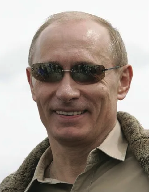 A man with light skin, short light hair, wearing dark sunglasses, smiling, dressed in a beige shirt and a brown jacket over his shoulder, outdoors with an overcast sky in the background.
