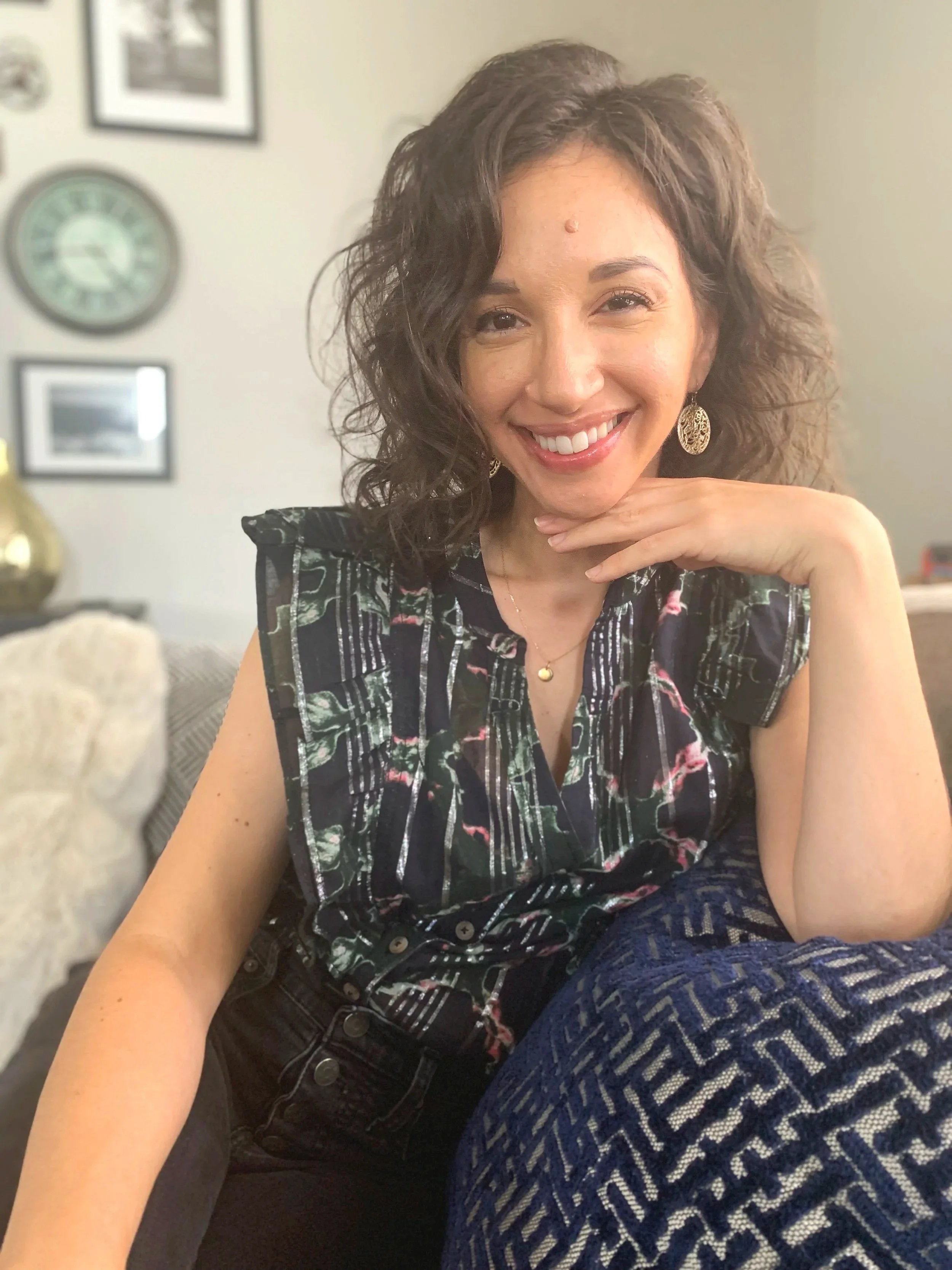 A woman with curly brown hair smiling and resting her chin on her hand, wearing a sleeveless patterned top and gold jewelry, sitting on a couch in a living room with framed pictures and a clock on the wall.
