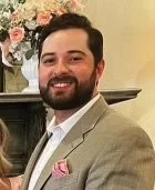 A man with a beard and dark hair smiling, dressed in a light-colored blazer, at a formal event.