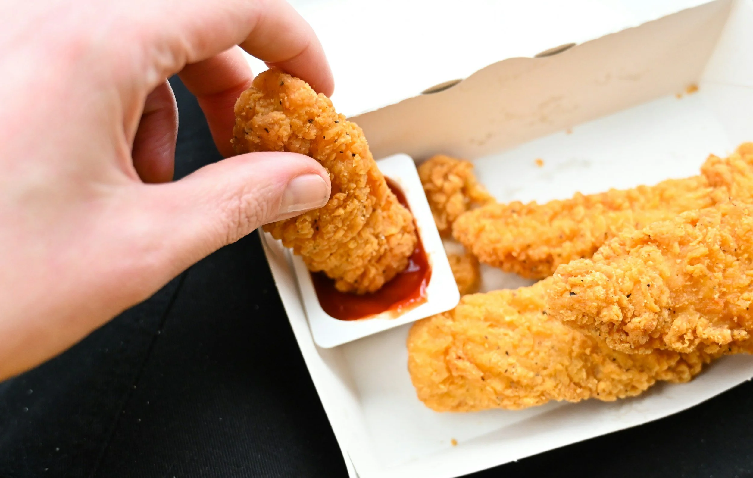 Close-up of hand dipping a piece of crispy fried chicken into a container of red dipping sauce, with more fried chicken on a tray in the background.