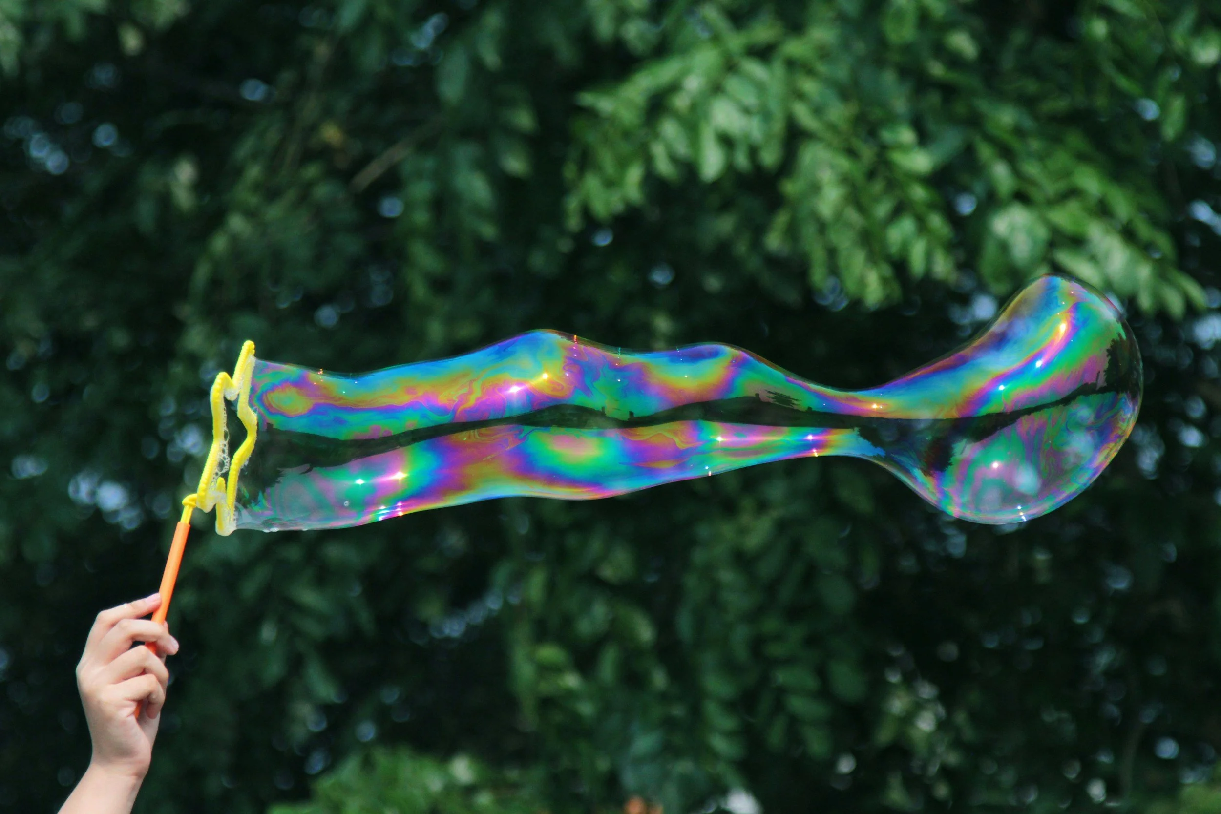 A person is holding a waving bubble wand creating a large, iridescent soap bubble outdoors with green trees in the background.