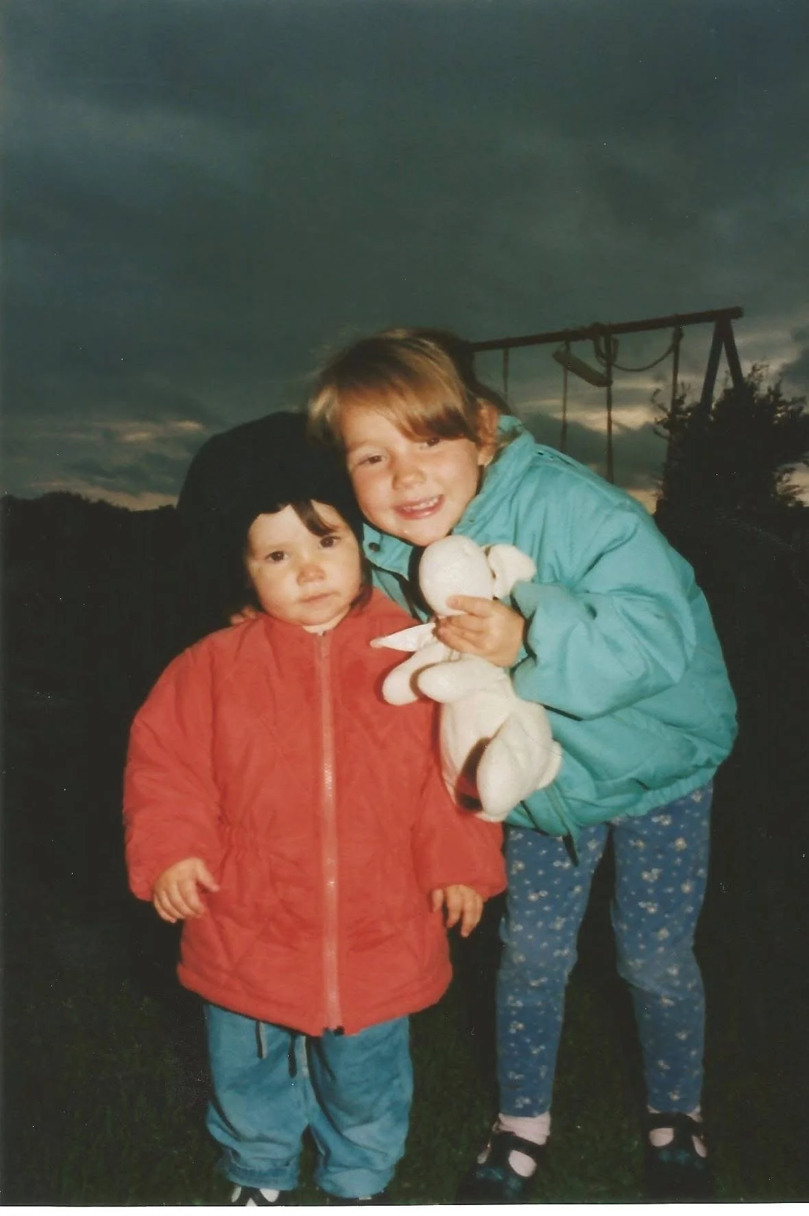 Two young girls, one with black hair and the other with blonde hair, standing outdoors during dusk or evening. The blonde girl is smiling and holding a stuffed animal, while the other girl is looking at the camera with a neutral expression. They are wearing jackets and pants.