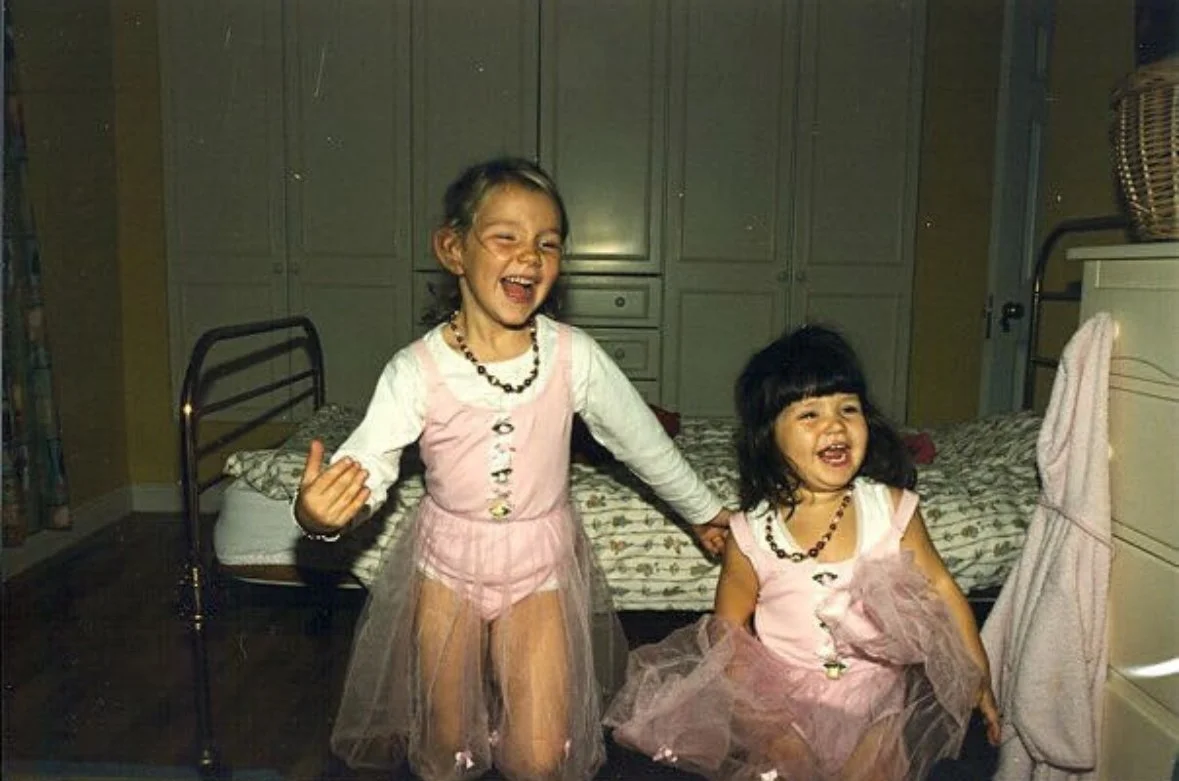 Two young girls in pink dresses and necklaces, laughing and holding hands in a bedroom with a bed and closet in the background.