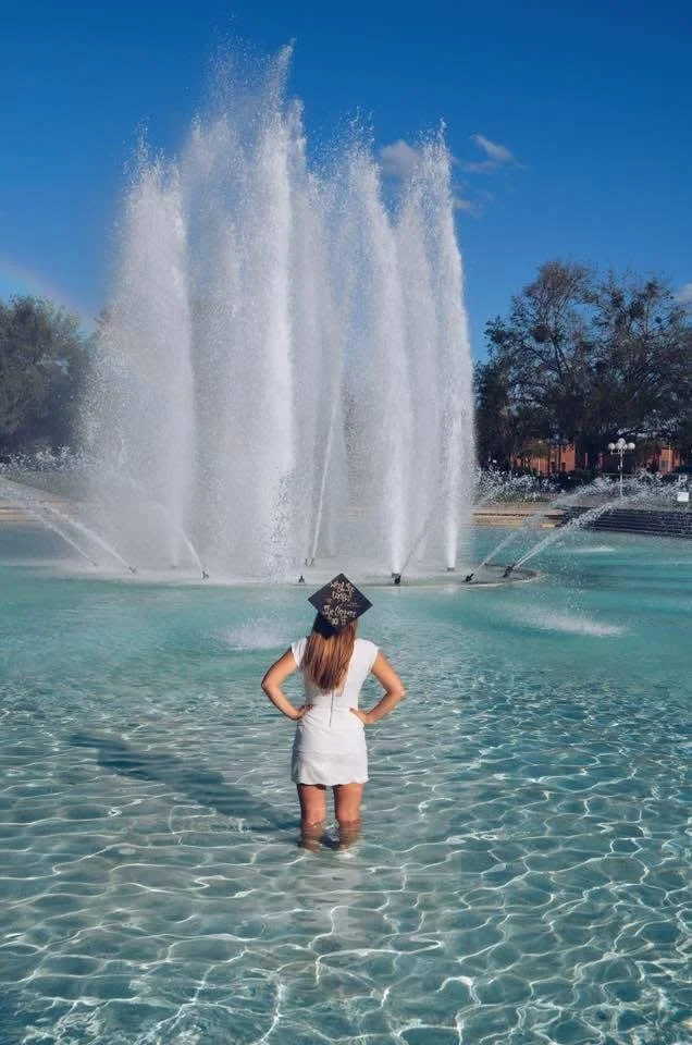A woman in a white dress standing in a shallow fountain pool, facing a large water fountain spray against a blue sky backdrop.