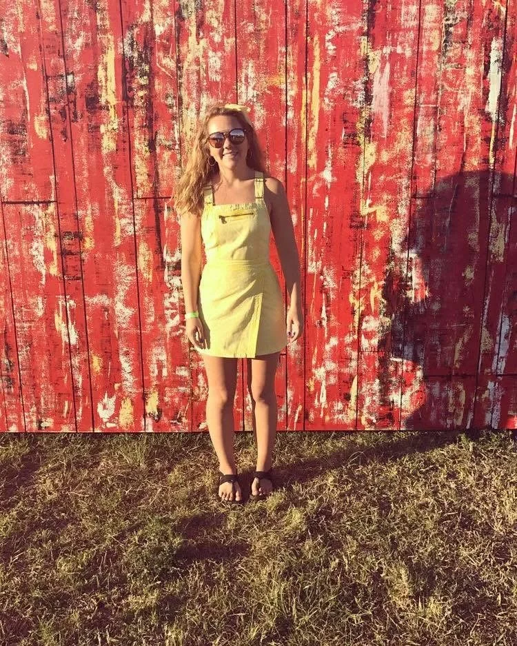 A woman with long, wavy blonde hair wearing sunglasses, a light yellow dress with shoulder cutouts, and black sandals standing on grass in front of a weathered red wooden wall.