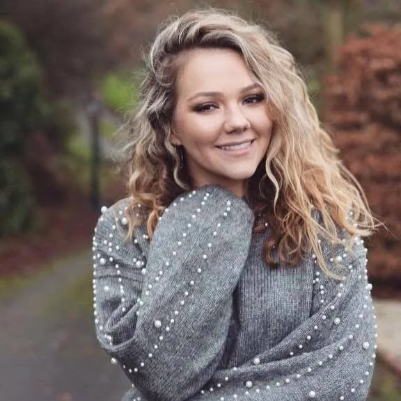 Young woman with curly blonde hair smiling outdoors in autumn, wearing a gray sweater with pearl embellishments.