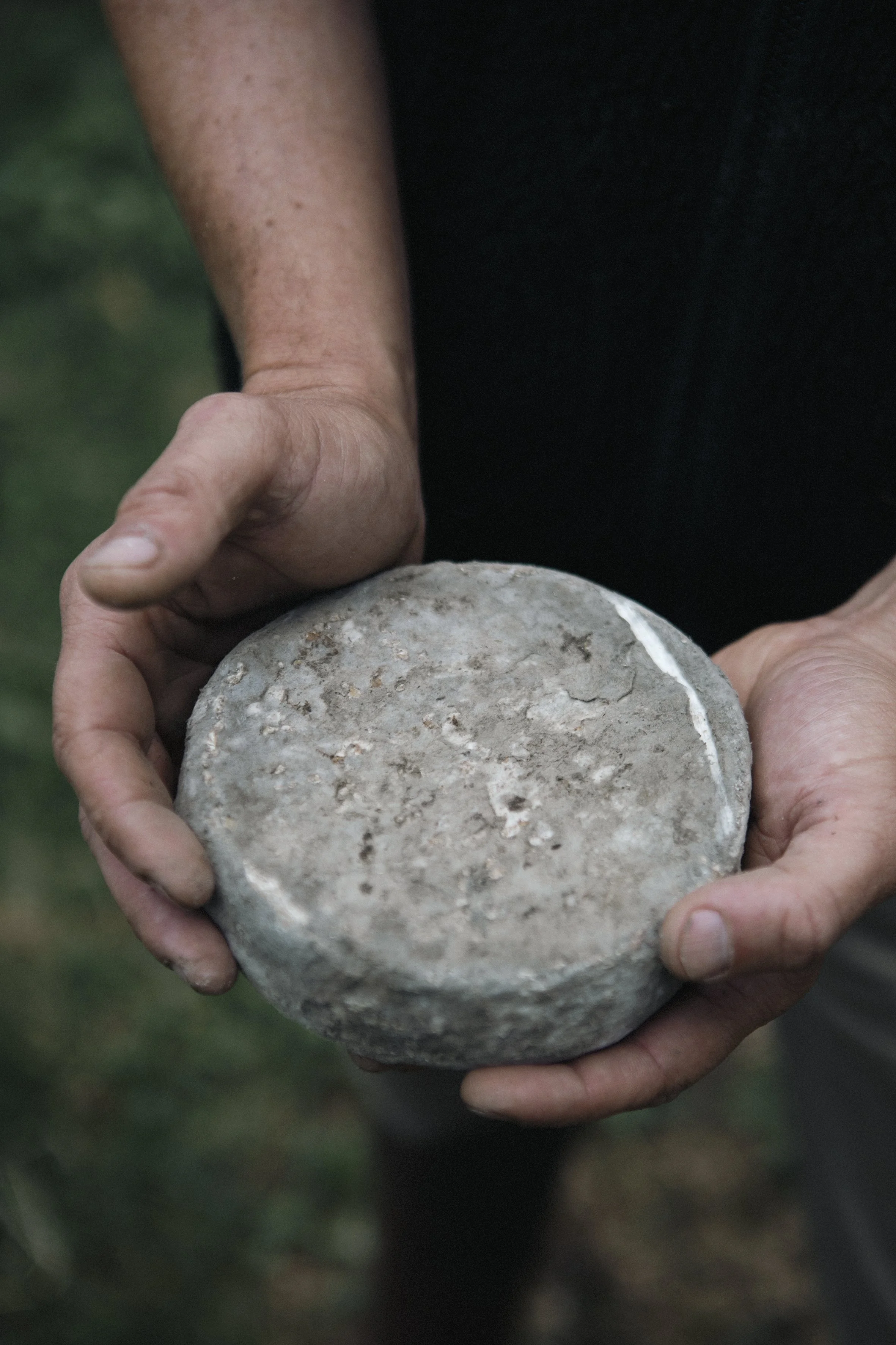 Une personne tient un objet circulaire en pierre ou en céramique, de couleur grise, avec une surface rugueuse et une bordure légèrement irrégulière.