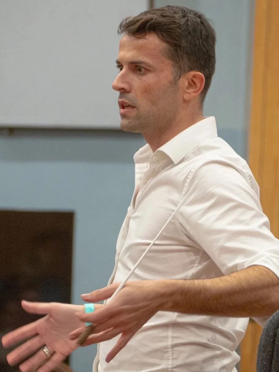 A man with brown hair, wearing a white shirt, standing in profile, gesturing with his hands, in a room with blue and wooden walls.