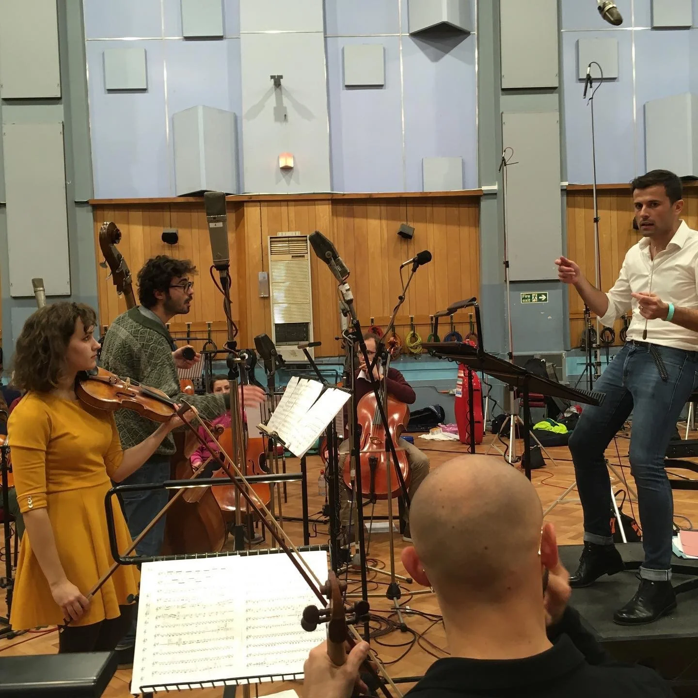 Group of musicians practicing in a recording studio with string instruments, microphones, and music stands, with a conductor guiding them.