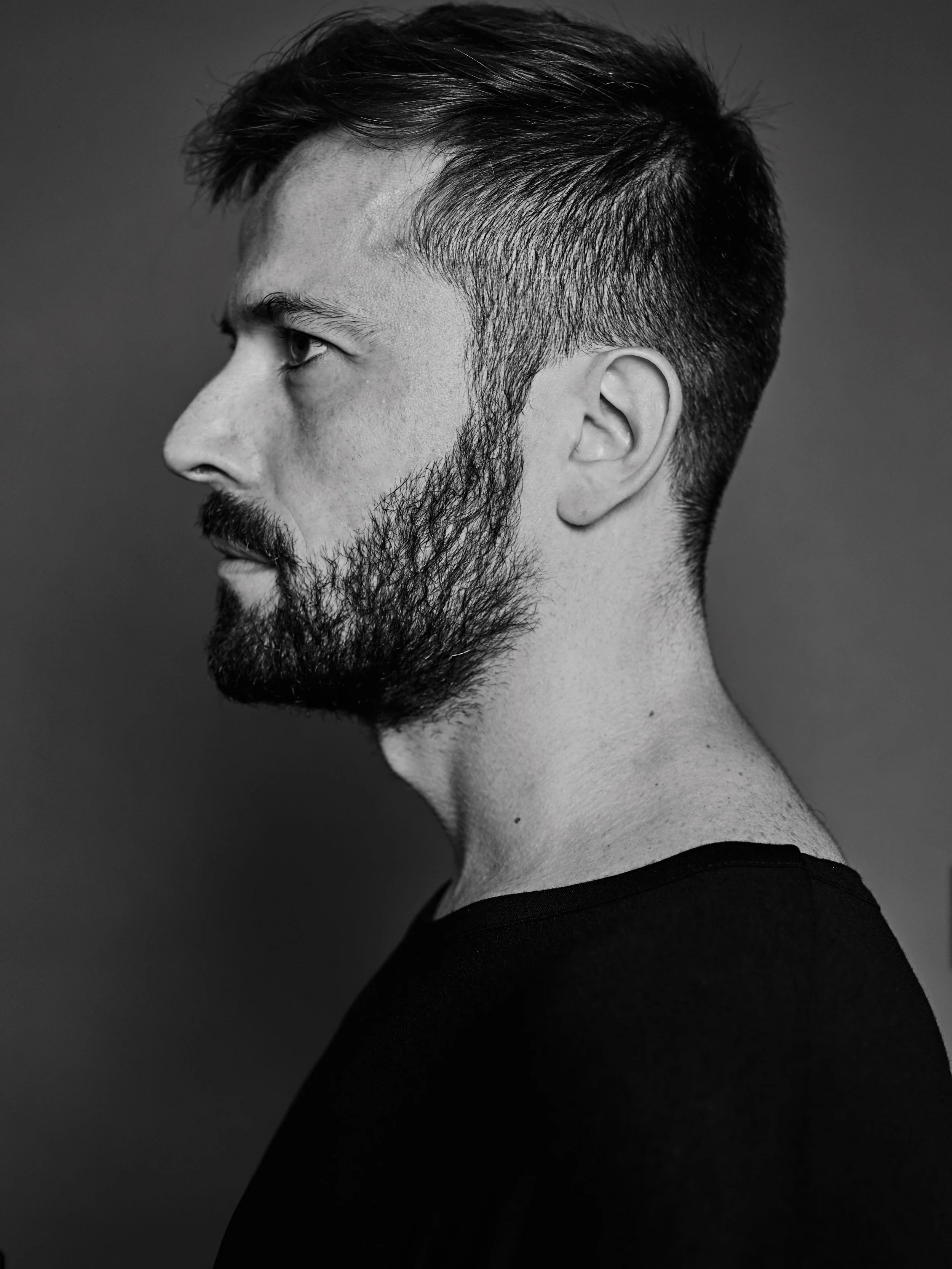 Black and white profile portrait of a young man with short hair and a beard, wearing a dark shirt, against a neutral background.