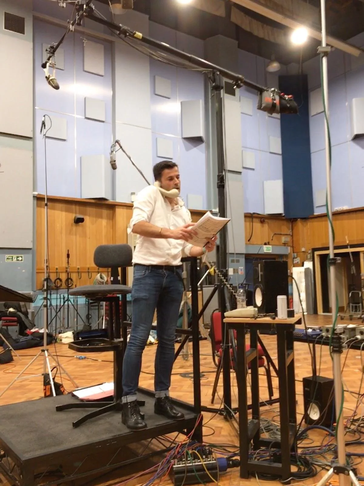 A person standing on a treadmill in a recording studio, holding a script, with audio equipment and cables around.