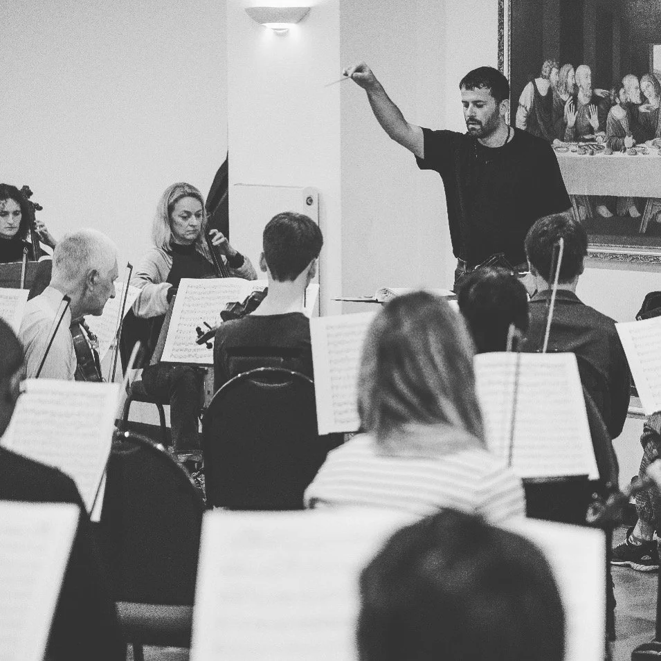 A man conducting an orchestra during a rehearsal, with musicians playing string instruments, in a room with a painting of The Last Supper on the wall.