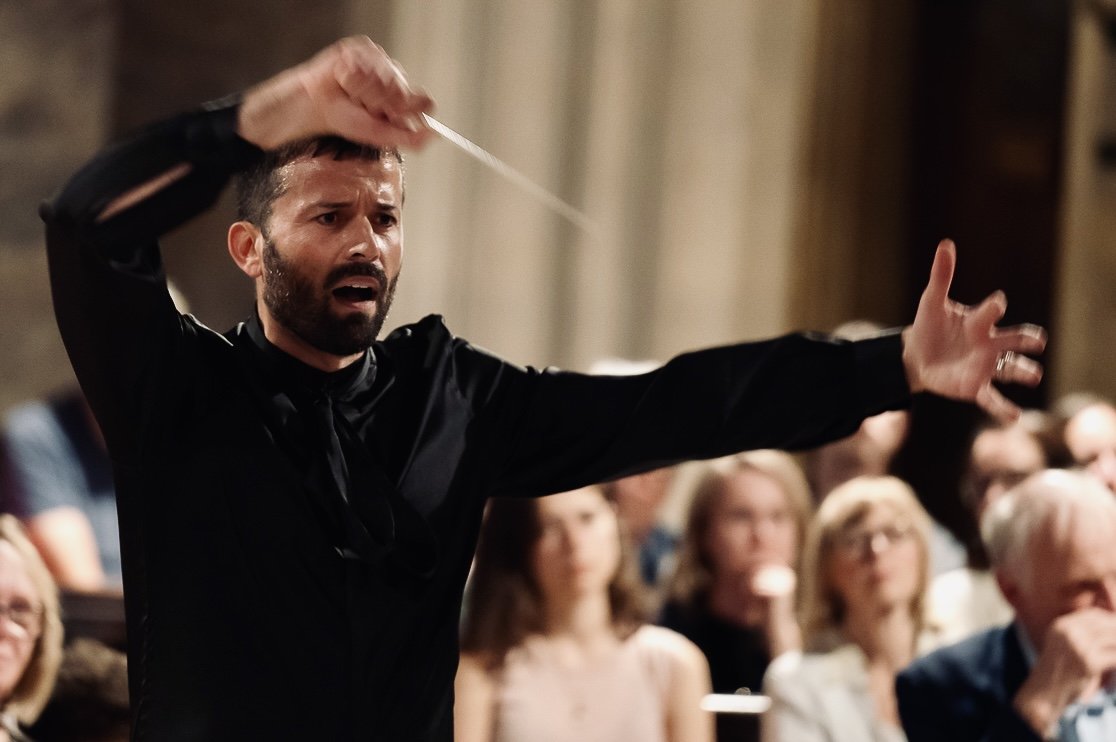 A man conducting an orchestra or choir, gesturing with his arm extended, in front of an audience.