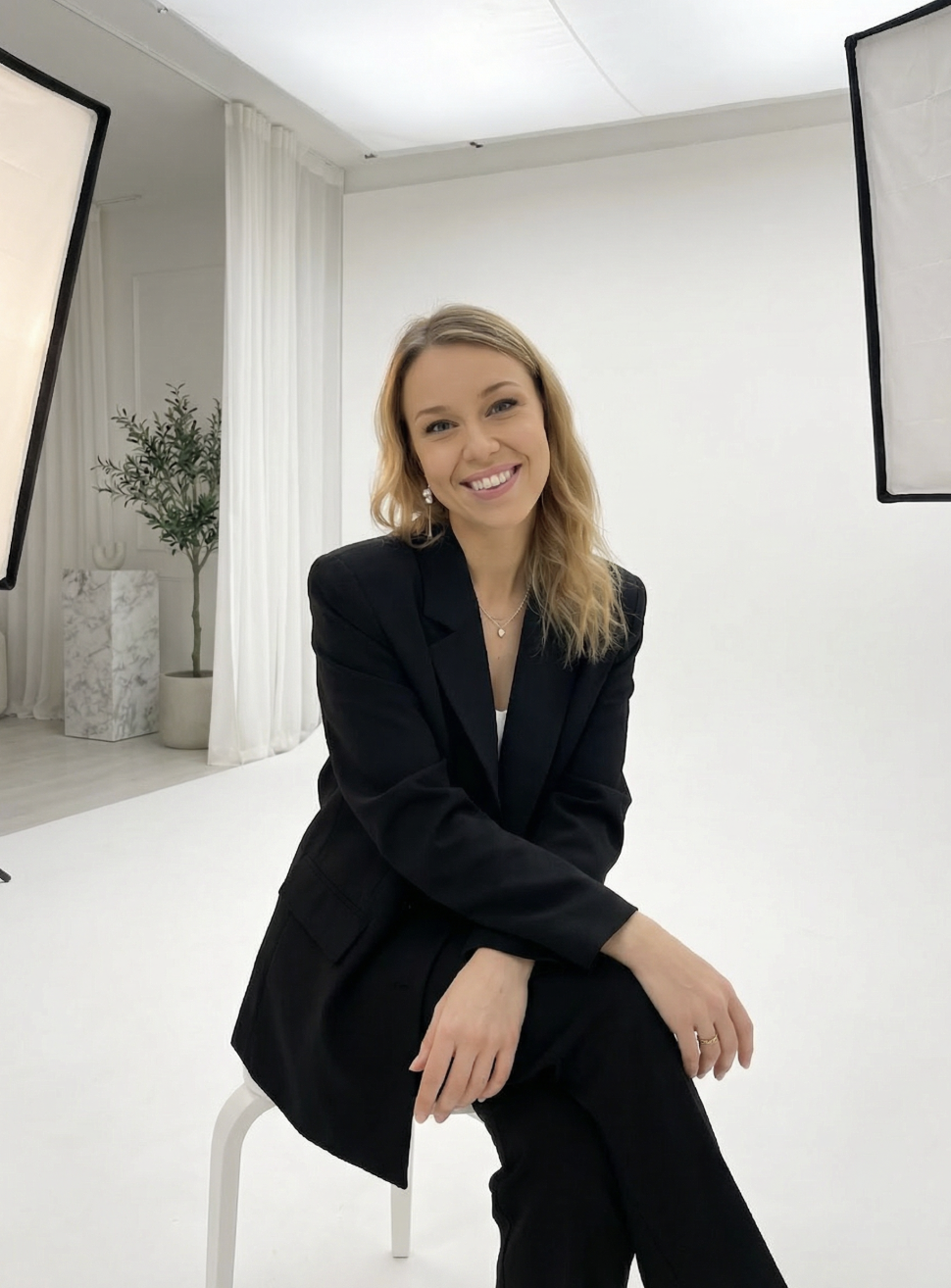 A woman sitting and smiling in a photography studio with white walls, soft lighting, and studio equipment visible.