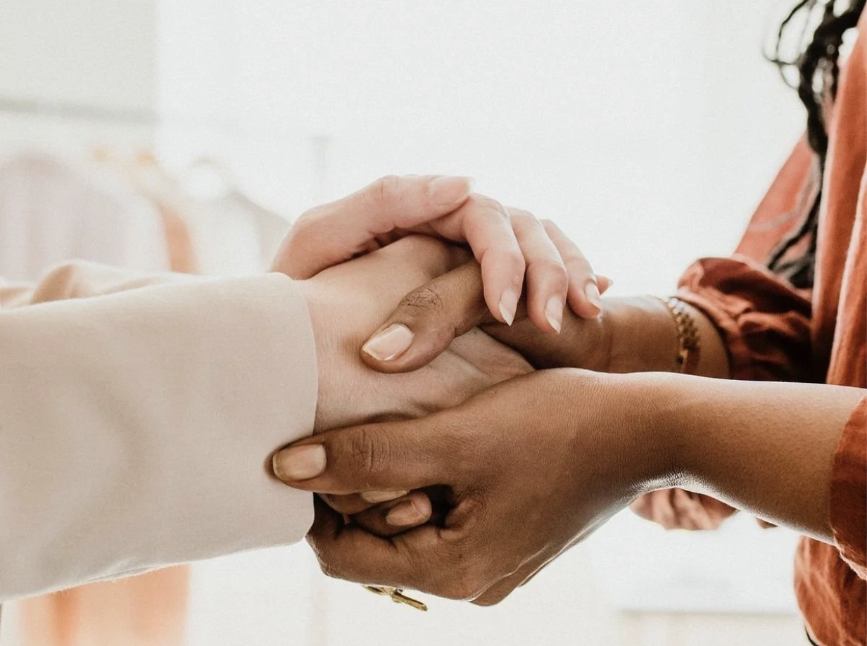 Multiple people holding hands together, symbolizing unity or support, with a plain indoor background.