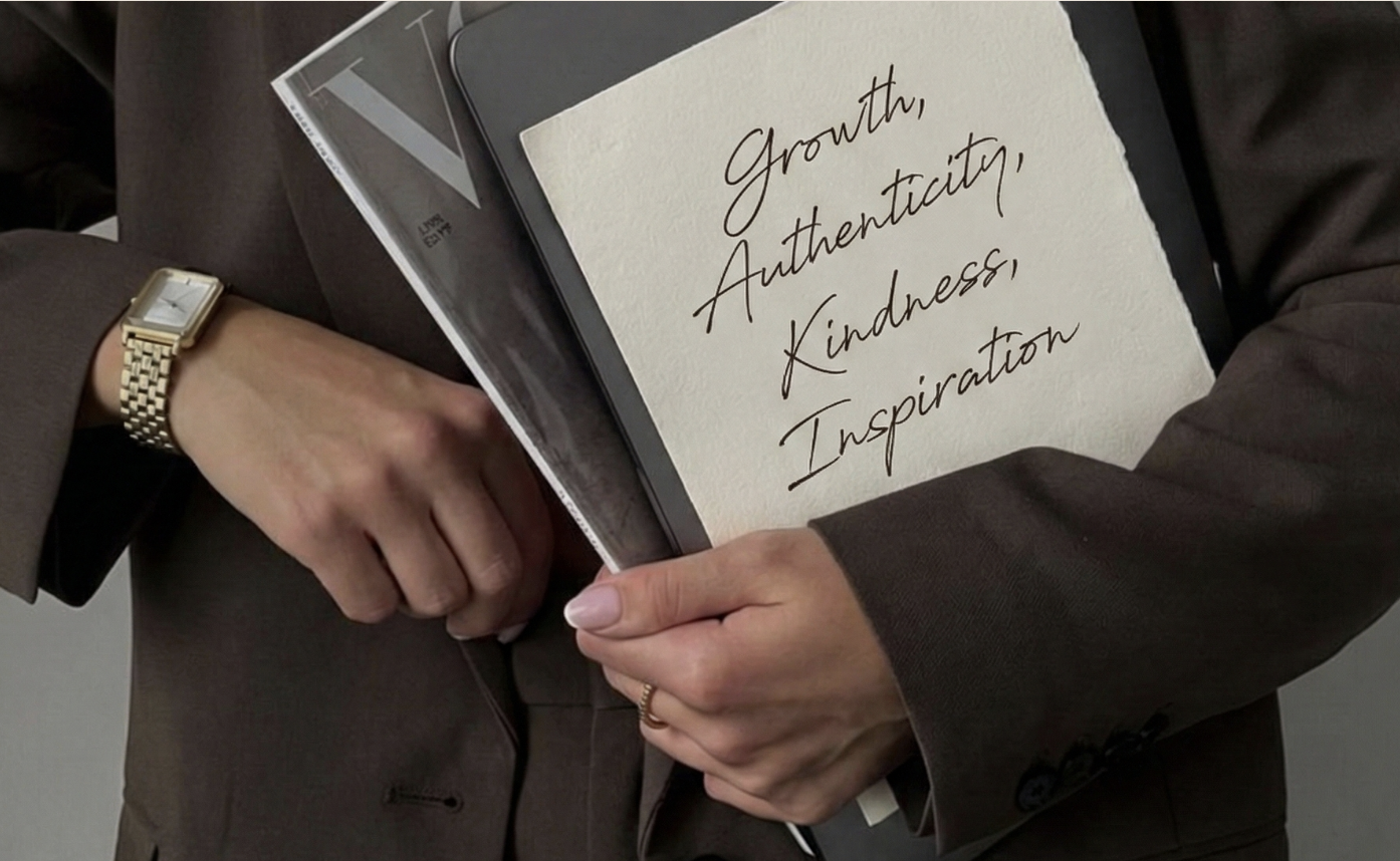 Close-up of a person holding a notebook with a handwritten note that says, "Growth, Authenticity, Kindness, Inspiration." The person is wearing a brown blazer and a gold watch, and is holding a smartphone.