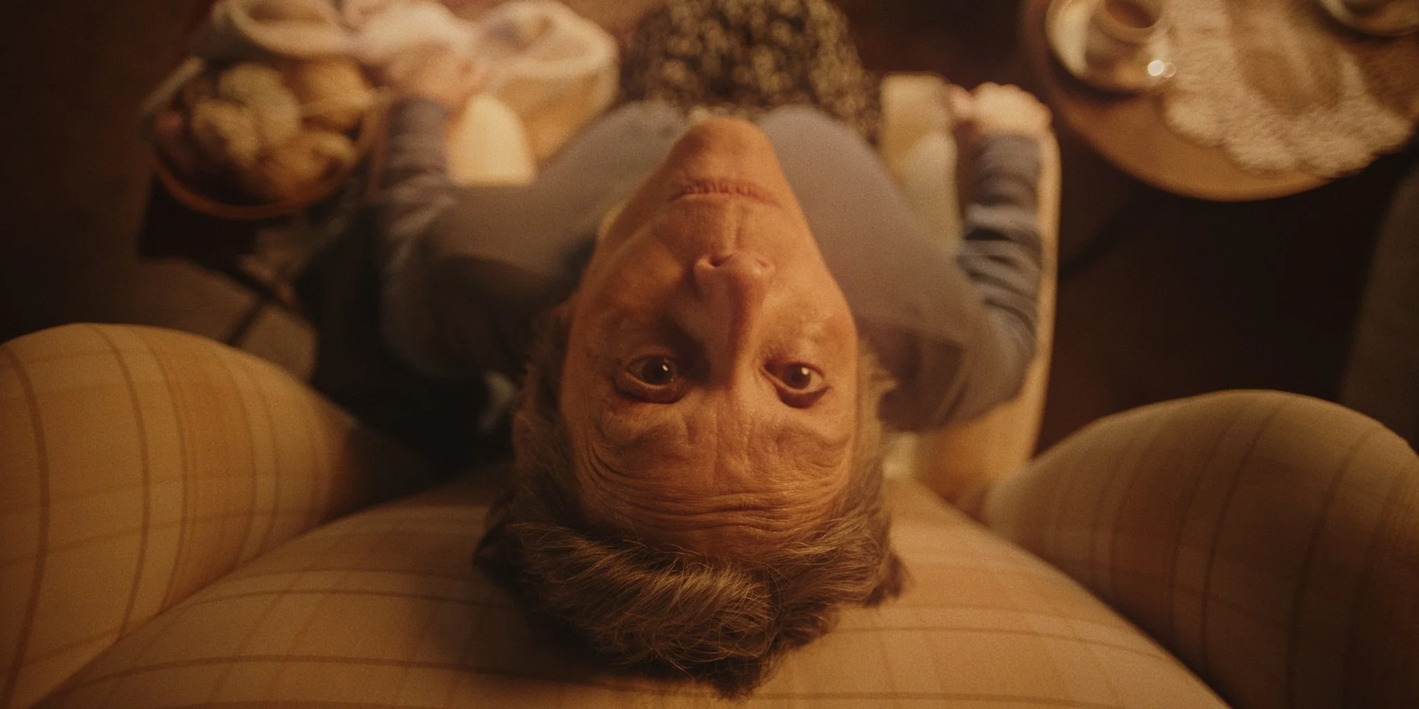 High angle, an elderly woman sits on a chair looking up at the camera, surrounded by cushions and in an indoor care home setting. Short film 'The Humbug' directed by Cole Paviour.