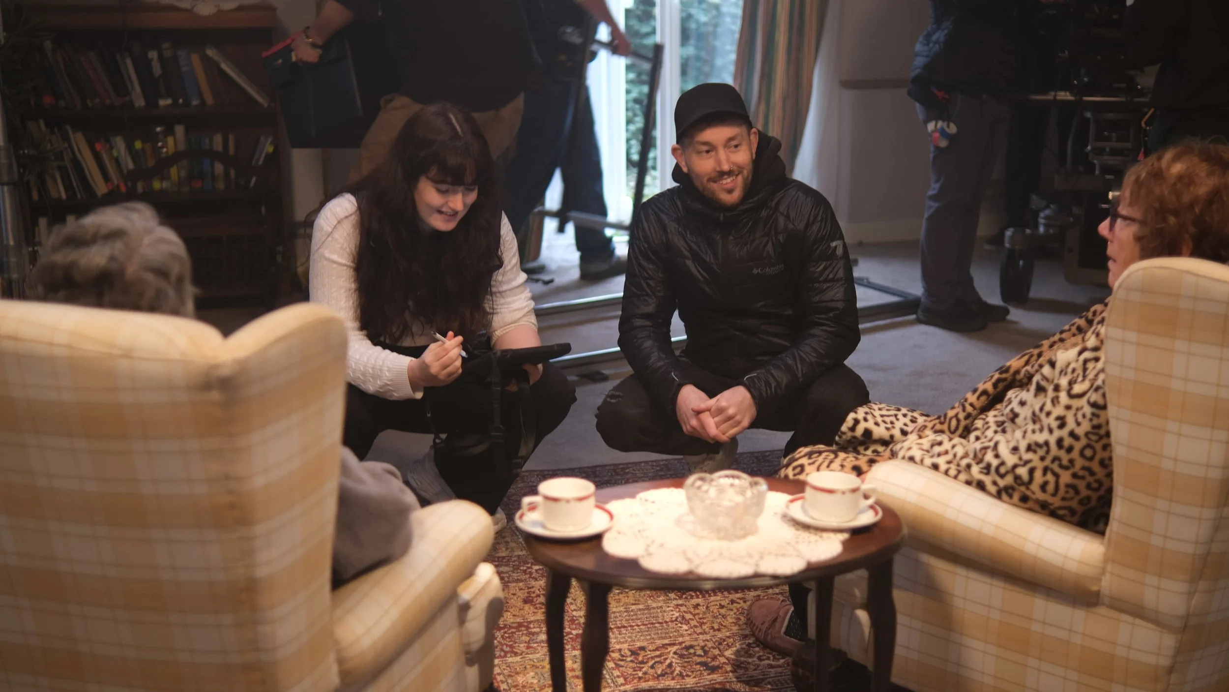 Director Cole Paviour speaks with actors on set, between takes. Two women are visible, one with dark hair and the other with red hair, and a man with a beard and a black jacket.
