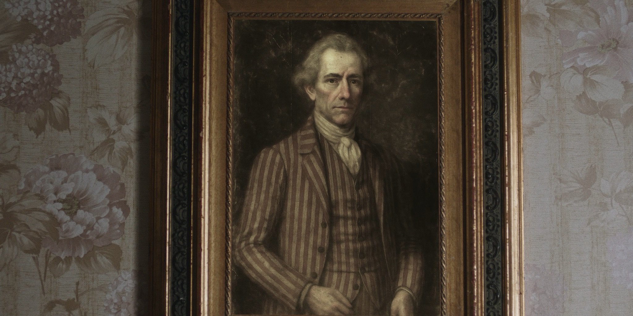 Portrait of a man with long gray hair, wearing a striped brown jacket, white shirt, and cravat, standing with arms crossed, framed and hanging on a floral-patterned wall.
