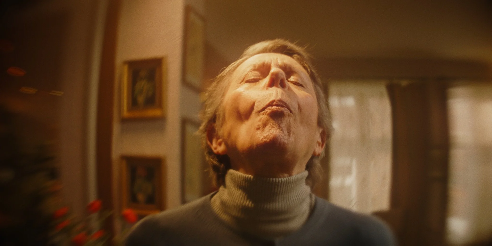 A woman with short gray hair and closed eyes wearing a turtleneck sweater, standing in a warm-lit room with framed pictures on the wall.