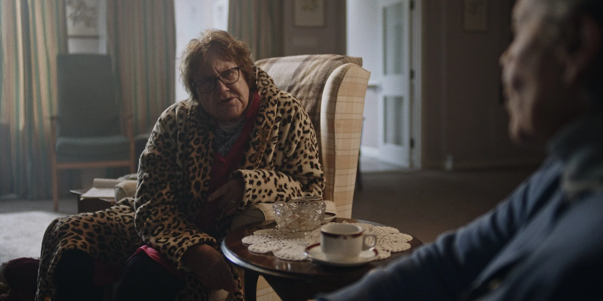 An elderly woman wearing glasses and a leopard print robe sitting and talking to a person with braided hair, with a small table between them holding a bowl, a cup, and a lace doily, in a cozy, well-lit room.