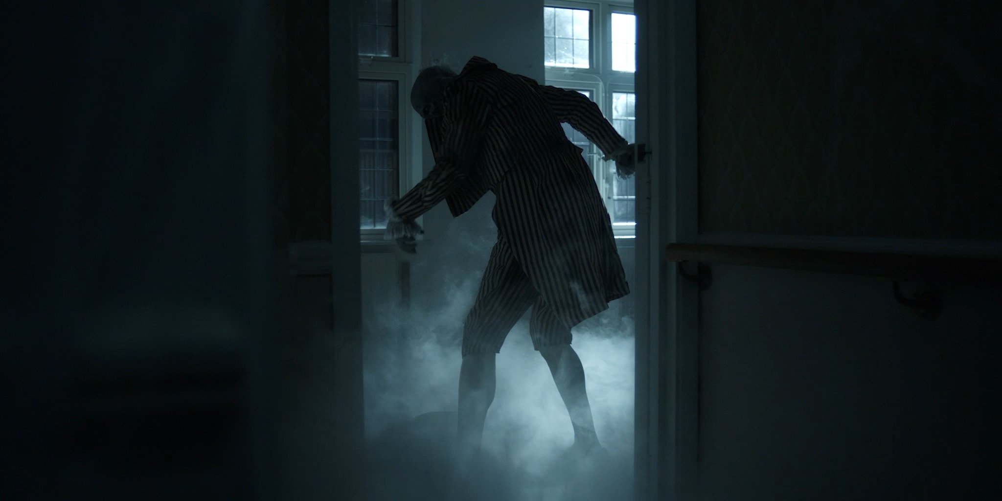 A person in striped pajamas standing in a dimly lit hallway filled with smoke or fog, holding a cloth, near a door with multiple glass-paneled windows, inside a house with pale blue walls and a dark corridor.