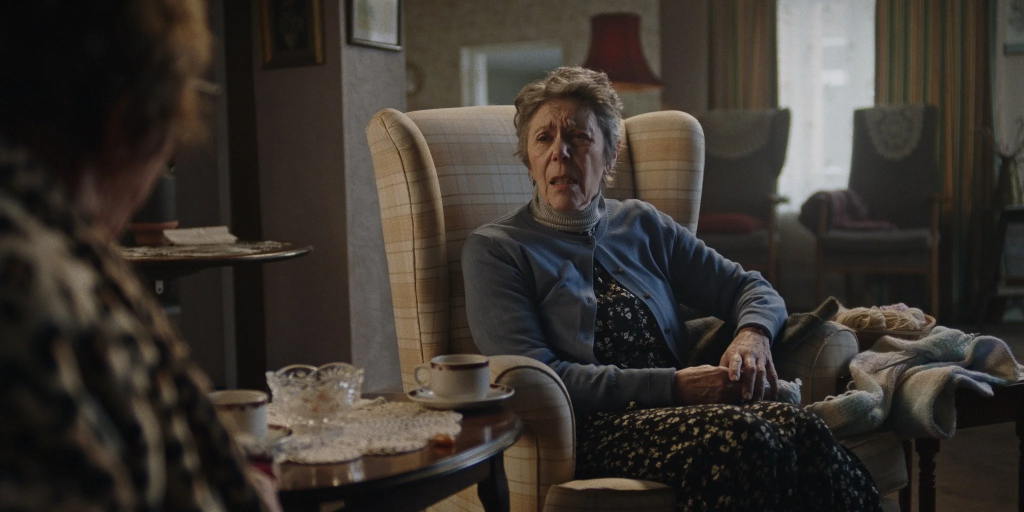 An elderly woman with short curly hair, wearing a grey cardigan over a black floral dress, sitting in a beige armchair, looking concerned or confused during a conversation with another person in a cozy living room.