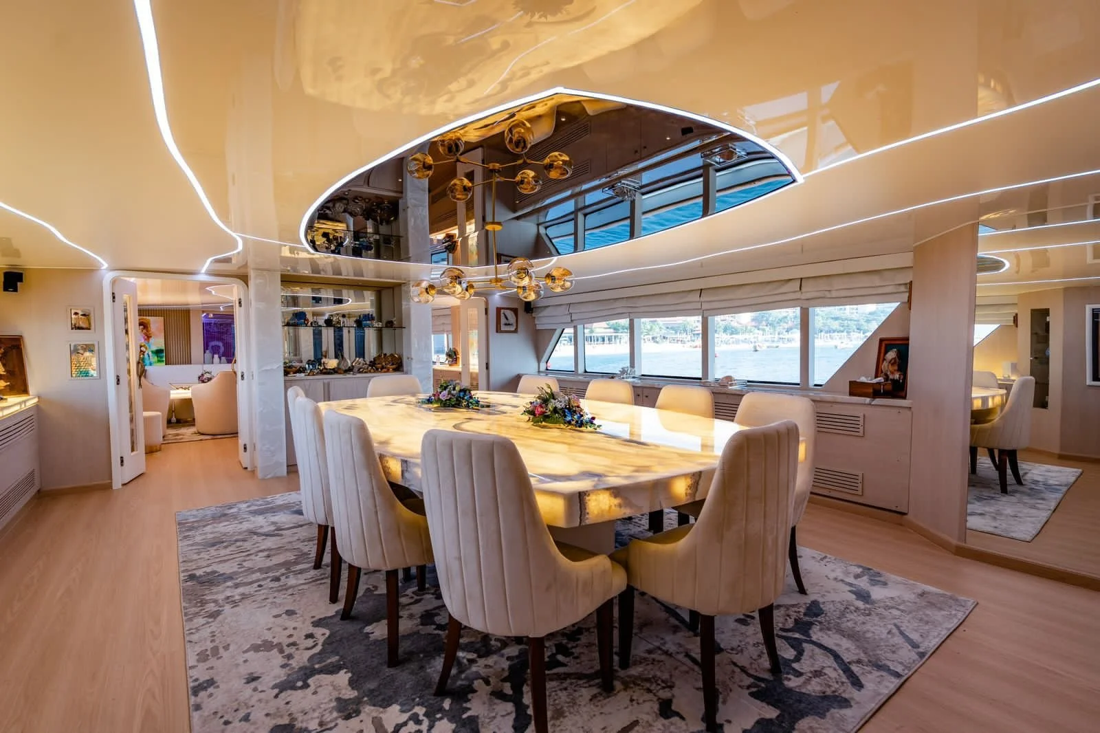 A luxurious dining room with a large oval marble table, surrounded by cream upholstered chairs, under modern lighting fixtures, with large windows overlooking a waterway, and decorative art displayed on the walls.
