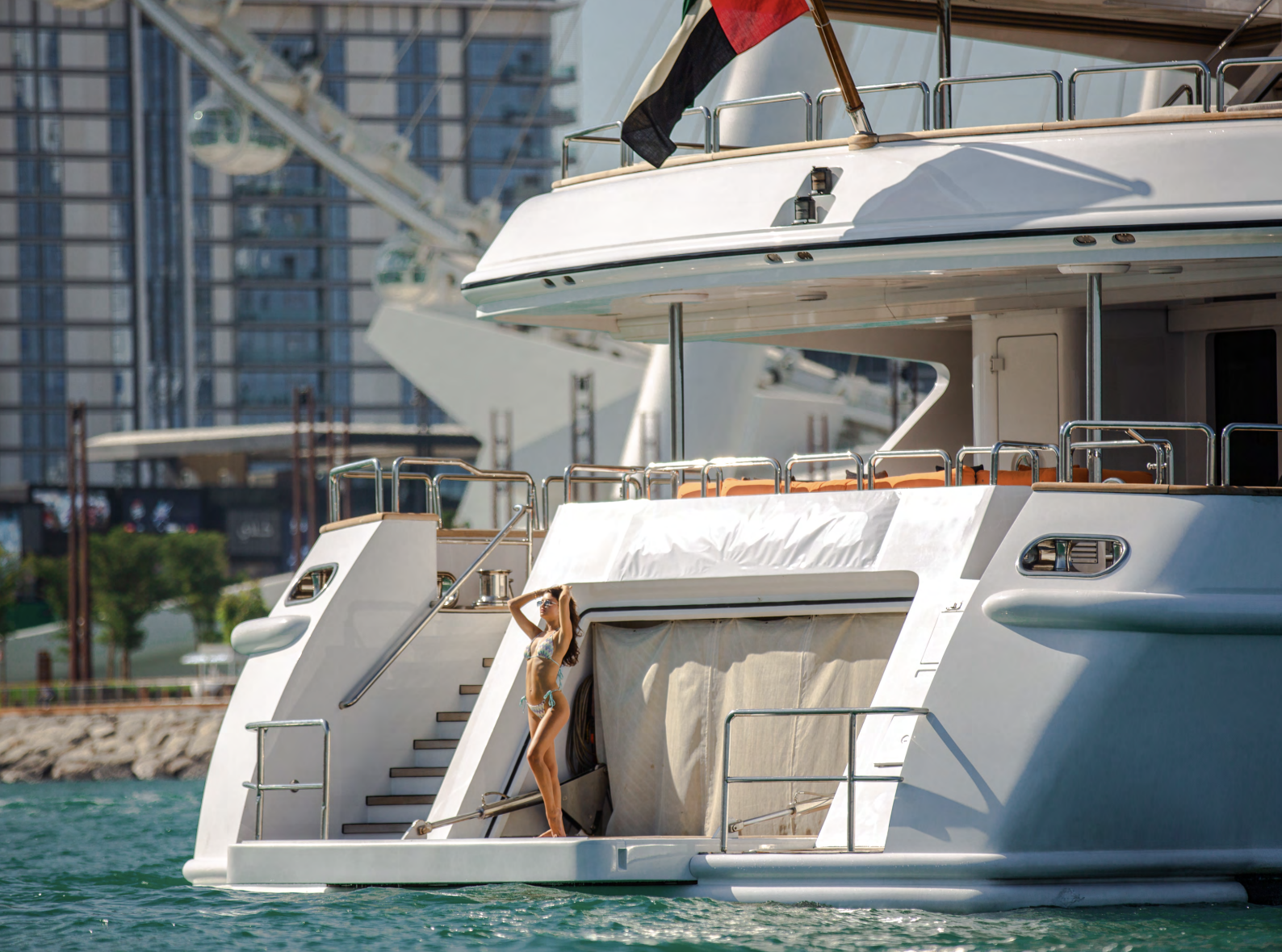 A woman in bikini posing on the back of a luxury yacht docked in a marina, with city buildings in the background.
