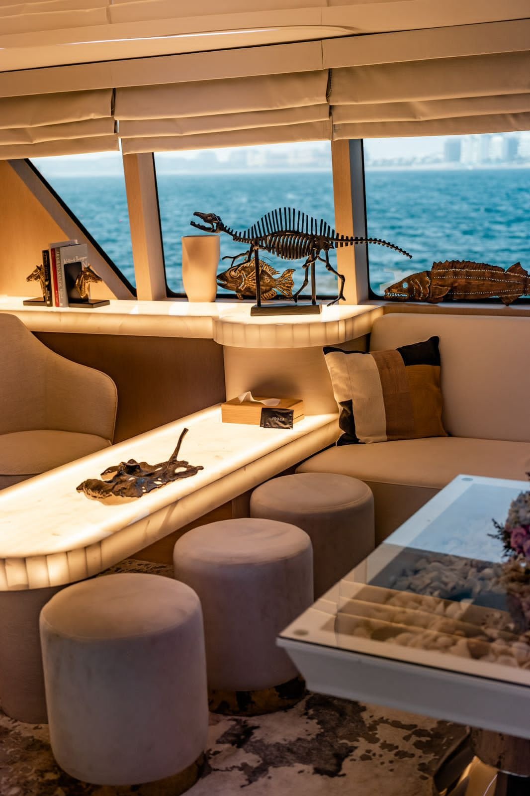 Interior room with a window view of water, displaying decorative dinosaur models, including a stegosaurus and a fish, on a ledge, with a sofa, pillows, stools, and a coffee table in the foreground.