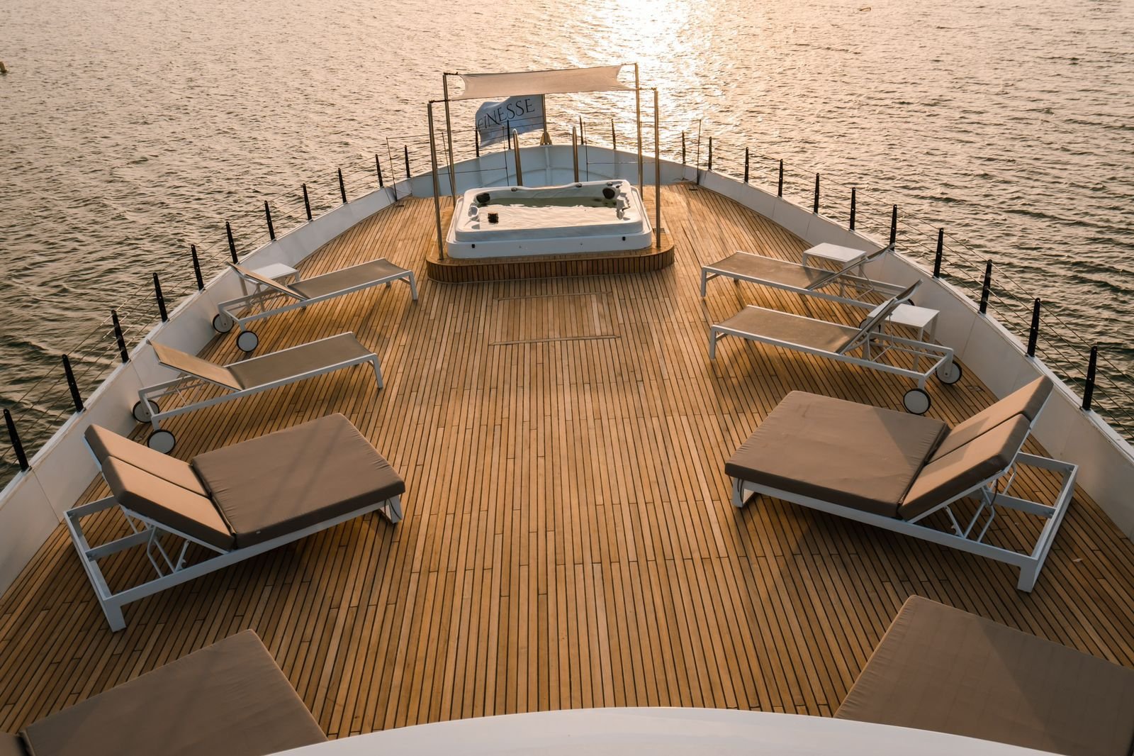 A deck of a boat with lounge chairs and a hot tub, overlooking the water at sunset.