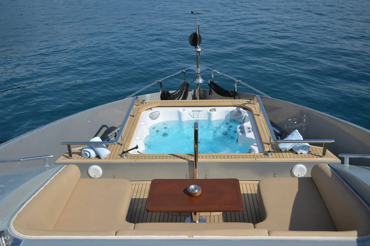 View from the rear of a yacht with a hot tub, beige cushioned seats, a wooden table, and towels, on calm blue water.