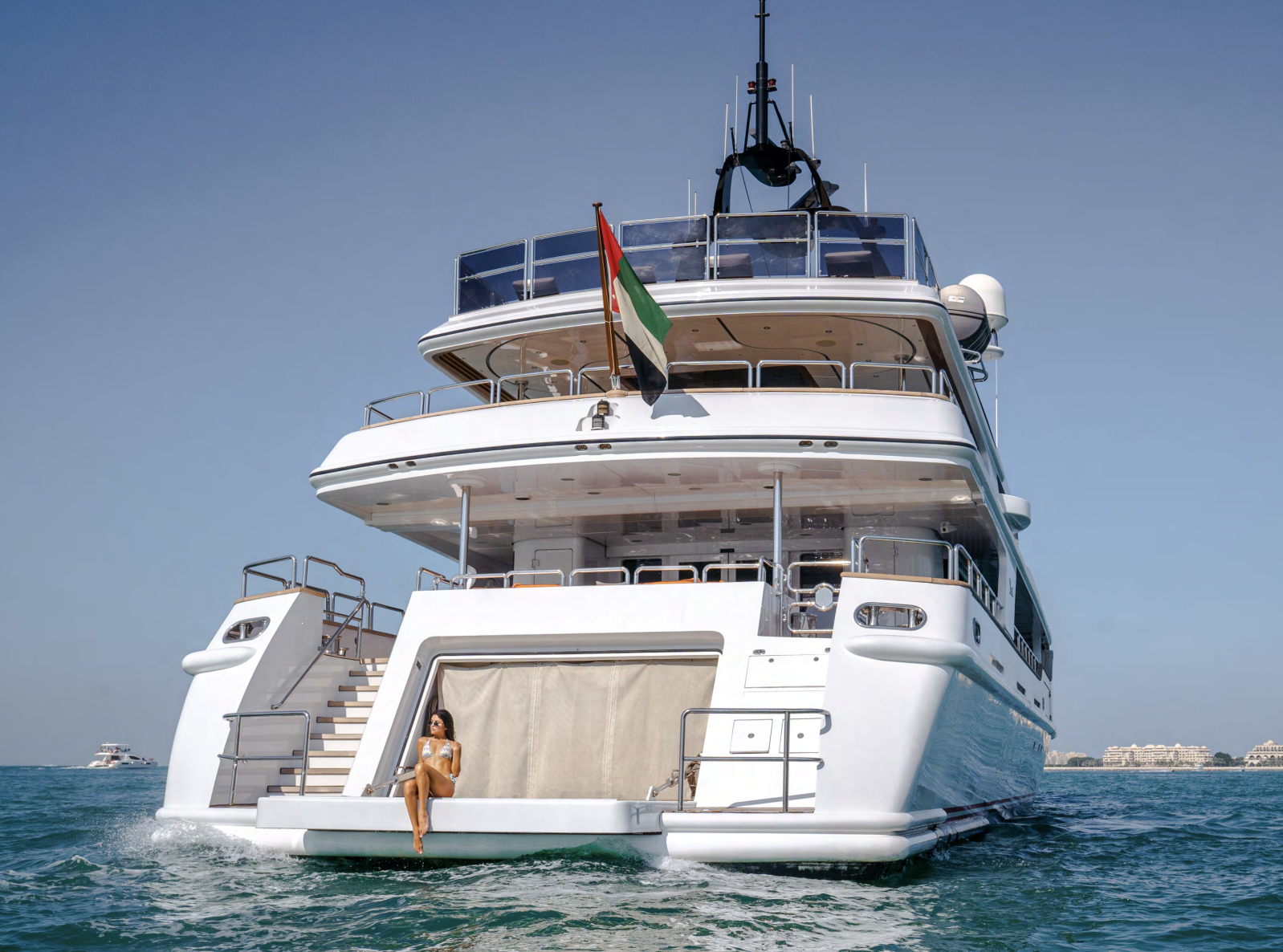 Luxury white yacht on the water with woman sitting at the stern, a boat in the distance, and buildings on the horizon under a clear blue sky.