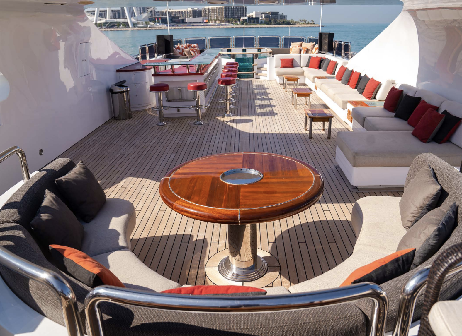 The luxurious outdoor deck of a yacht with seating, tables, a bar area, and a view of the water and city skyline in the background.