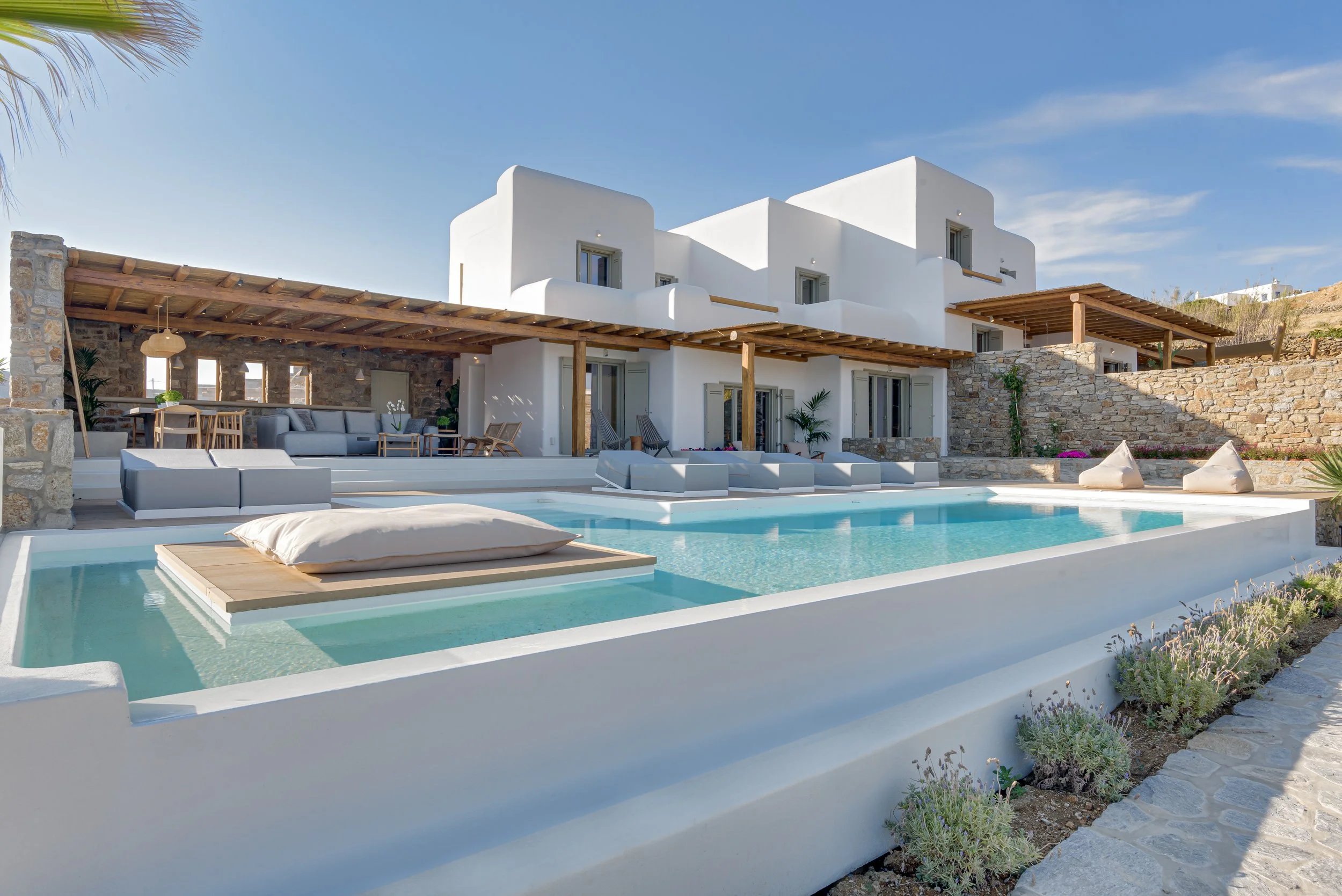 A modern white villa with stone accents, outdoor pool with floating cushions, covered patio with seating and dining areas, and a clear blue sky.